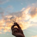A hand holds a light bulb against a scenic sunset sky, symbolizing creativity and innovation.
