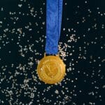 Close-up of a shimmering gold medal with blue ribbon on a black surface, symbolizing victory and achievement.